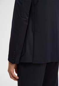 Vista posteriore di una persona che indossa una giacca e pantaloni da abito blu navy scuro, con la manica della giacca che mostra quattro bottoni neri vicino al polso.