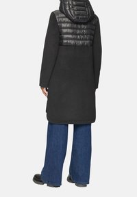 Abrigo negro con sección superior acolchada y capucha, combinado con pantalones de denim de pierna ancha y botas negras. Textura suave, diseño elegante.