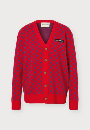 Femme portant un cardigan rouge à pois bleus, une chemise blanche, un pantalon large beige et des chaussures noires pointues, debout face à l’avant.