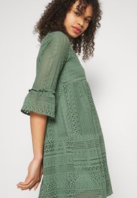 Green lace dress featuring structured embroidery, a round neckline, three-quarter sleeves with ruffles, and a flared fit that falls above the knee.