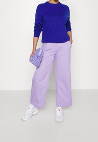 Lila tröja, lavendelfärgade vida byxor och en mjuk lavendelfärgad axelväska. Vita sneakers med detaljer. Släta material och avslappnad passform.