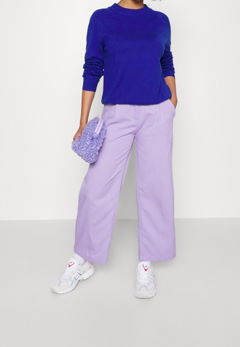 Lila tröja, lavendelfärgade vida byxor och en mjuk lavendelfärgad axelväska. Vita sneakers med detaljer. Släta material och avslappnad passform.