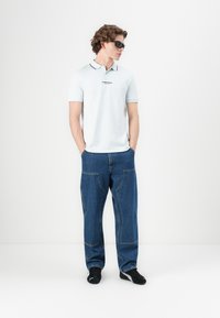 Svetlomodré polo tričko s čiernymi akcentmi, kombinované s voľnými, vysokým pásom tmavými rifľami. Modelka má na sebe čierne slnečné okuliare a tenisky.