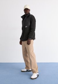 Chaqueta de forro polar negra con cuello con cremallera y panel de goma, pantalones beiges, zapatillas blancas con detalles negros y una gorra beige.