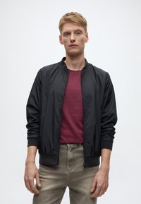 Juoda bombonešio striukė su užtrauktuku priekyje, su ribotomis rankogaliais ir apykakle, dėvima ant bordo spalvos marškinėlių ir šviesiai pilkų džinsų. Lygi audinio tekstūra.