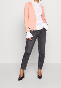 Blazer pêche à l'avant ouvert, blouse blanche texturée avec des poignets à volants, jean foncé et chaussures noires à talons hauts.