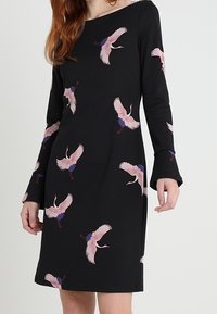 Robe noire à manches longues ornée de motifs colorés de grues, avec un col rond et des manches évasées, fabriquée dans un tissu doux et extensible.