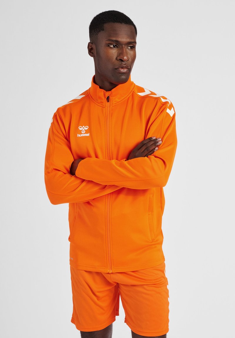 Oranje trainingsjas en shorts met witte schouderstrepen en logo. Gemaakt van glad, lichtgewicht materiaal, met een rits aan de voorkant en zakken.