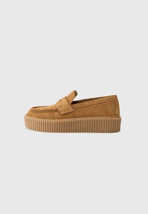 Brun ruskind slip-on loafer med tyk ribbet gummisål og en dekorativ rem over toppen, vist i sideprofil på en lys baggrund.