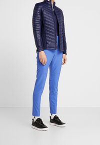 Marinblå vadderad jacka med hög krage, blå sportbyxor och svarta sneakers. Jackan har sidosömmar; byxorna har en slät yta.