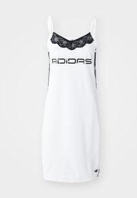 Bela atletska obleka z črnimi čipkastimi robovi, ki ima v odebeljenem tisku logotip "ADIDAS" in kontrastne črne črte ob straneh.