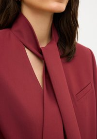 Burgundy blus med knytning vid halsen, med en V-formad utskärning och en enda bröstficka. Slät tyg med en matt yta.