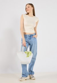 Hvid tote taske med grøn mønstret sløjfe, stofhåndtag og en struktureret form. Model iført en cremefarvet top og lyseblå jeans, stående på en lys baggrund.