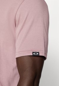 Chemise à manches courtes en coton rose clair à texture uni. Présente une étiquette noire avec un logo blanc au bas de la manche.