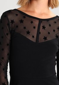 Czarna odzież z długimi rękawami; półprzezroczysta warstwa w fakturze czarnych gwiazd; dopasowana talia; okrągły dekolt.