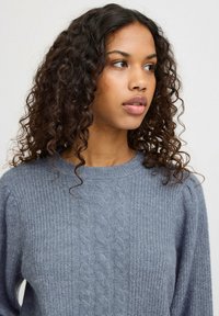 Pull en tricot gris avec un motif en torsades, un col côtelé et une texture douce. Manches légèrement amples.