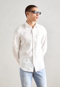 Weißes Leinenhemd mit Knopfleiste und einer Tasche, kombiniert mit hellblauen, verwaschenen Jeans. Trägt dunkle, rechteckige Sonnenbrillen.