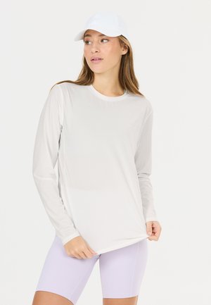 Femme portant une chemise blanche à manches longues, une casquette blanche et un short de cycliste violet clair, regardant sur le côté tout en tenant l'ourlet de la chemise.