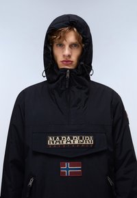 Black anorak jacket with a zippered neck, featuring a large front pocket, "Napapijri" logo, and a Norwegian flag patch. Soft texture.