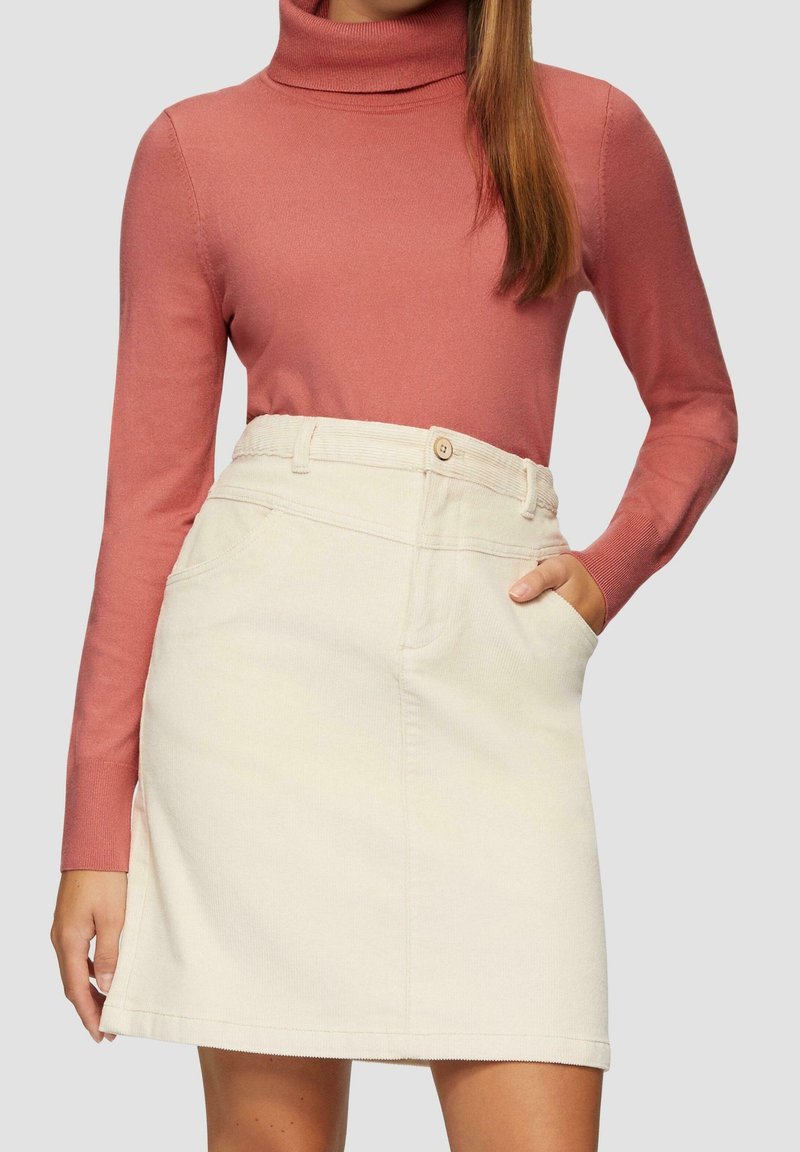Femme portant un pull à col roulé rose poussiéreux et une jupe en velours côtelé crème avec des poches, une main dans la poche, sur un fond uni.