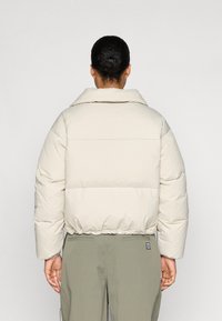 Pufferjack in lichtbeige, met een hoge kraag, oversized pasvorm en elastische onderrand. Gepaard met olijfgroene broek met subtiele zakken.