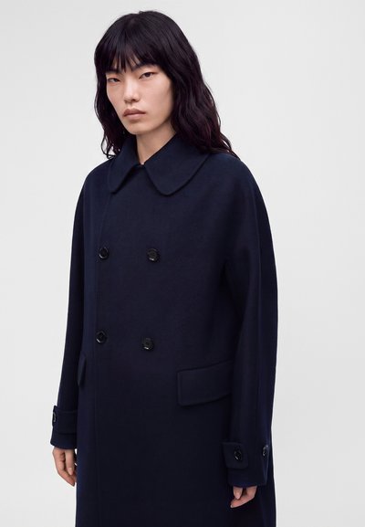 Femme aux longs cheveux noirs portant un manteau bleu marine foncé à double boutonnage avec un grand col et des poignets boutonnés sur un fond uni.