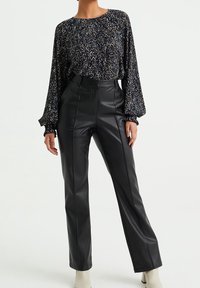 Blouse à manches longues, à motifs sombres avec des manches amples, associée à un pantalon en cuir noir taille haute avec des coutures latérales et des jambes droites.