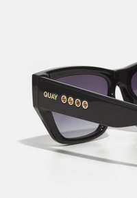 Black angular sunglasses with gradient purple lenses, featuring gold accents and four decorative studs on the side. Text reads "QUAY."