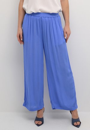 Kobieta ubrana w luźne, szerokie jasnoniebieskie spodnie, zestawione z ciemnoniebieskimi, odkrytymi na palcach butami na obcasie i białą wsuniętą bluzką.