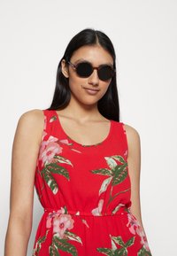 Vestido floral vermelho com flores rosa e folhas verdes, apresentando um decote em forma de sorriso e cintura elástica. Óculos de sol redondos e escuros com detalhes em tartaruga.