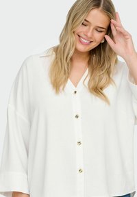Camicia bianca over-size con scollatura a V, maniche corte e bottoni dorati, realizzata in un tessuto testurizzato e con una vestibilità rilassata.