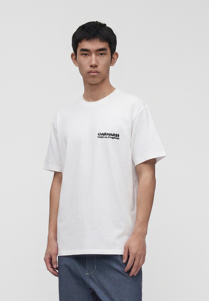 Giovane uomo che indossa una t-shirt bianca Carhartt con logo, in piedi davanti a uno sfondo grigio chiaro uniforme, guarda avanti.