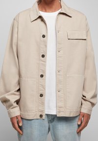 Beżowa kurtka zapinana na guziki z kieszenią na klatce piersiowej i dwiema dolnymi kieszeniami, noszona na białej koszulce i jasnoniebieskich dżinsach.