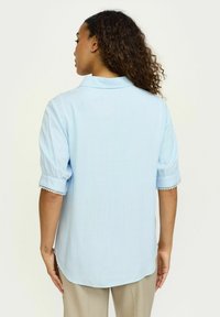 Sky blue shirt with rolled sleeves and decorative trim. Soft fabric, collared design, and curved hem at the back, shown from behind.