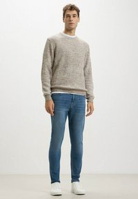 Maglione in maglia beige con texture a coste, scollatura a girocollo; abbinato a jeans blu slim-fit e sneakers bianche. Modello in piedi contro uno sfondo uniforme.