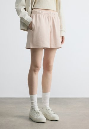 Personne portant un short beige clair à taille élastique, un haut beige, une veste légère, des chaussettes blanches et des baskets blanches, debout sur un sol gris.