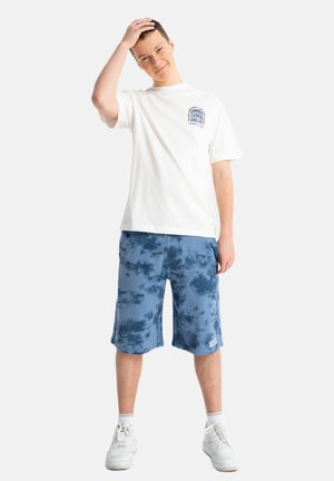 Nastolatek z krótkimi włosami, ubraną białą koszulkę z grafiką, niebieskie szorty tie-dye, białe skarpetki i białe trampki, stoi z jedną ręką na głowie.