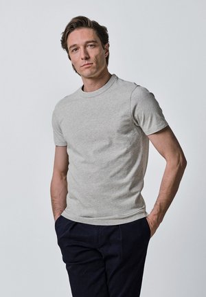 Jonge man met lichte huid en bruin haar draagt een lichtgrijs T-shirt met korte mouwen en donkere broek, staand met handen in de zakken.