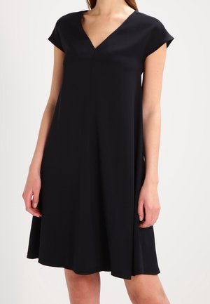 Vrouw draagt een zwarte jurk tot op de knie met korte mouwen en een V-hals, staand tegen een effen lichte achtergrond.