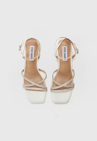 Sandali beige con tacco alto e punta quadrata, caratterizzati da fascette incrociate impreziosite da strass trasparenti e una fibbia alla caviglia su una superficie liscia.