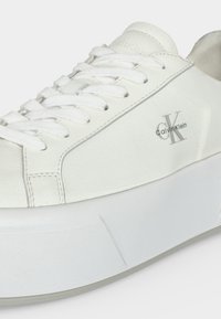 Λευκό χαμηλό παπούτσι Calvin Klein με παχιά σόλα, λευκά κορδόνια και μαύρο λογότυπο "CK Calvin Klein" στην πλαϊνή επιφάνεια.
