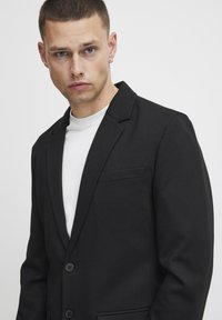 Black blazer with a notched lapel, single chest pocket, and two buttons. Worn over a white t-shirt, the fabric has a smooth texture.