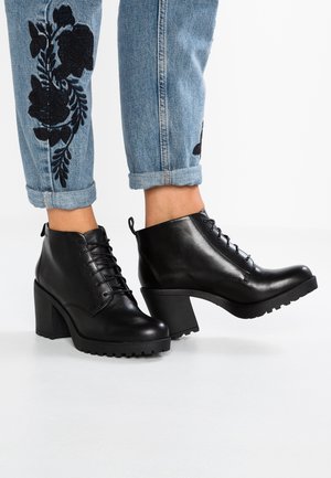 Bottines en cuir noir à lacets avec talons épais portées avec un jean bleu retroussé orné de broderies florales noires.