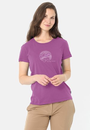 Giovane donna con capelli castani lunghi che indossa una maglietta viola Vaude con un disegno circolare di una bicicletta e un paesaggio e pantaloni beige su sfondo bianco.