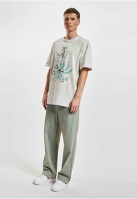 Šedé oversized tričko s tyrkysovým grafickým designem, spárované s světle zelenými širokými džínami a bílými teniskami.