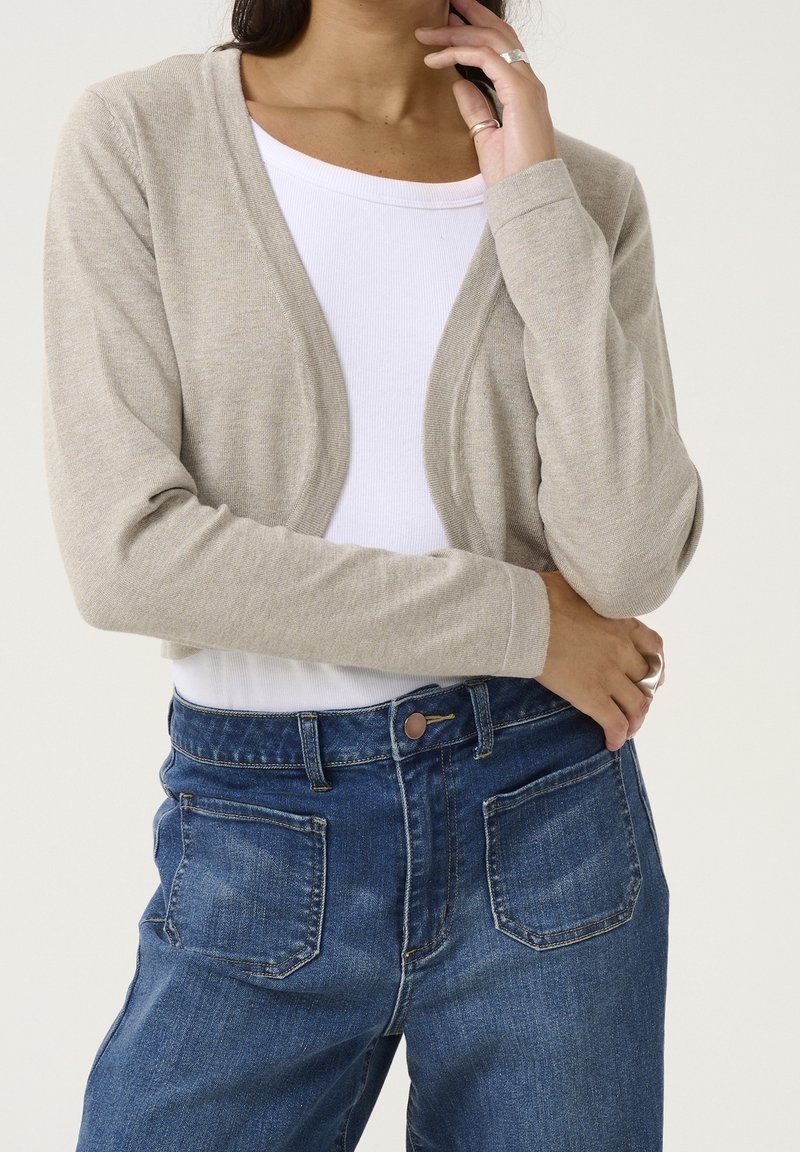 Femme portant un cardigan court beige sur un haut blanc et un jean bleu taille haute, une main touchant le visage et l'autre bras croisé.