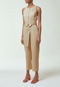 Femme portant une combinaison beige sans manches avec une ceinture à la taille et des sandales corail à talons avec brides, debout devant un fond clair uni.