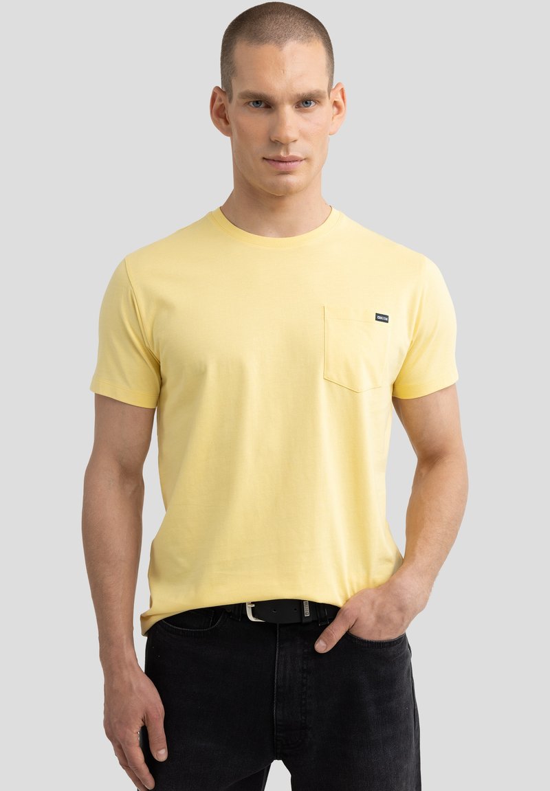 Geltonas medvilninis marškinėliai trumpomis rankovėmis, apvaliu iškirpimu, priekinė kišenė ir maža prekės ženklo logo ant krūtinės. Dėvimi su tamsiomis džinsais.