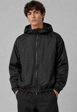 Giovane uomo che indossa una giacca nera con cappuccio e zip, con sottili motivi diagonali e mani in tasca, su sfondo grigio uniforme.