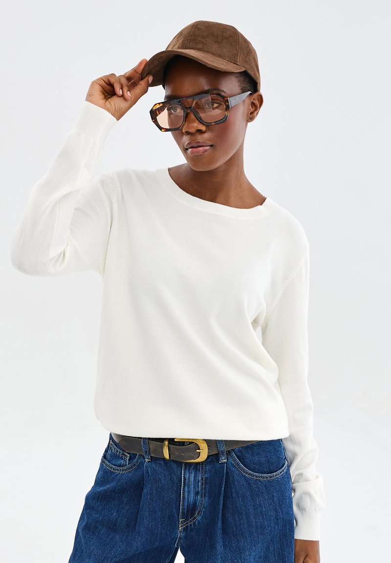 Persoon met oversized schildpadbril, wit langmouwen sweater, blauwe jeans met zwarte riem en bruine pet, die de pet aanpast.